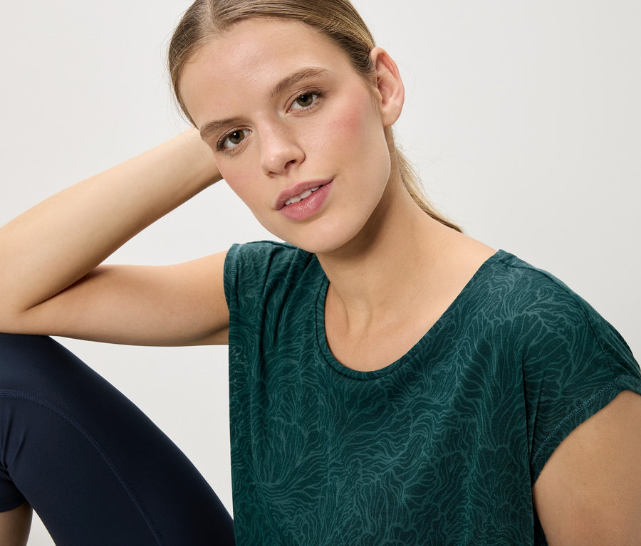 Eine Frau sitzt und trägt ein grünes Shirt mit Muster und blaue Leggings.