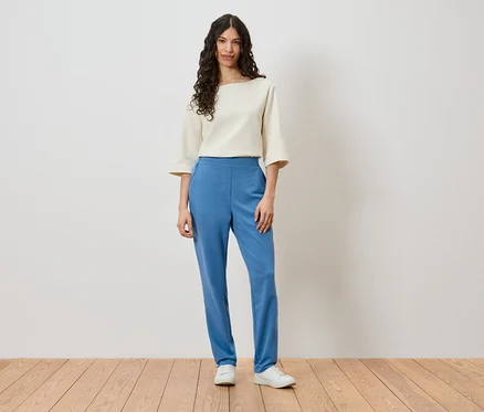 Eine Frau steht vor einer Wand und trägt ein cremefarbenes Oberteil, blaue Hosen und weiße Schuhe.