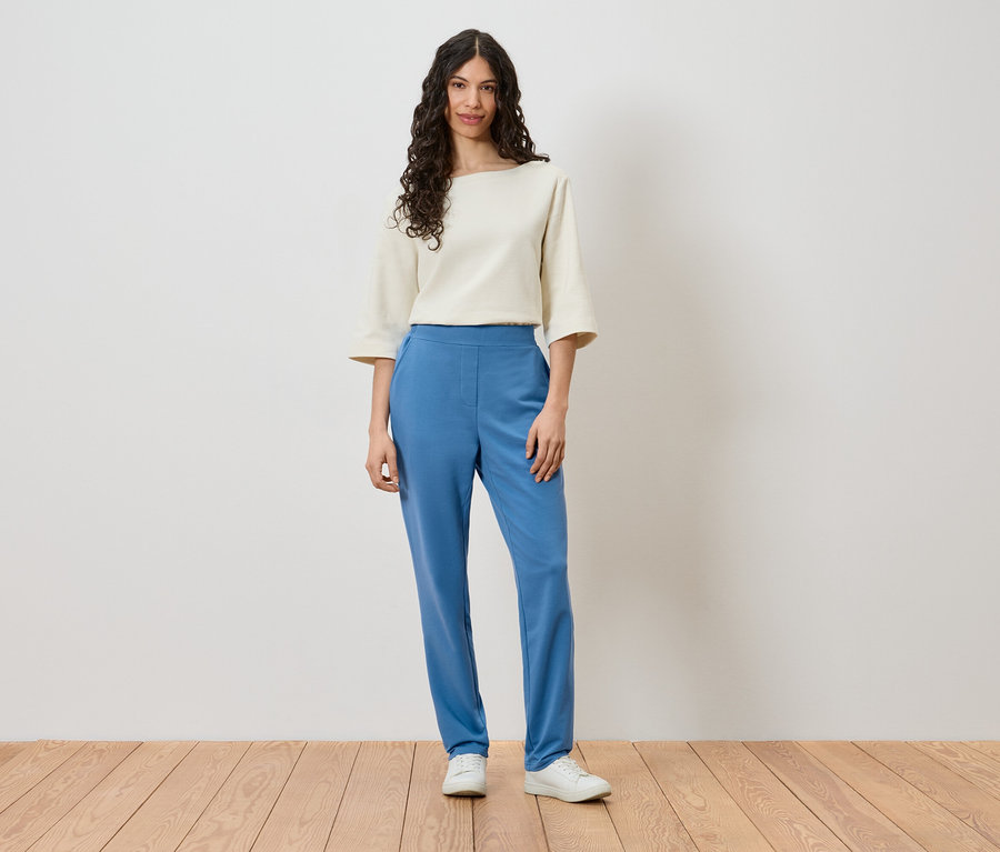 Eine Frau steht vor einer Wand und trägt ein cremefarbenes Oberteil, blaue Hosen und weiße Schuhe.