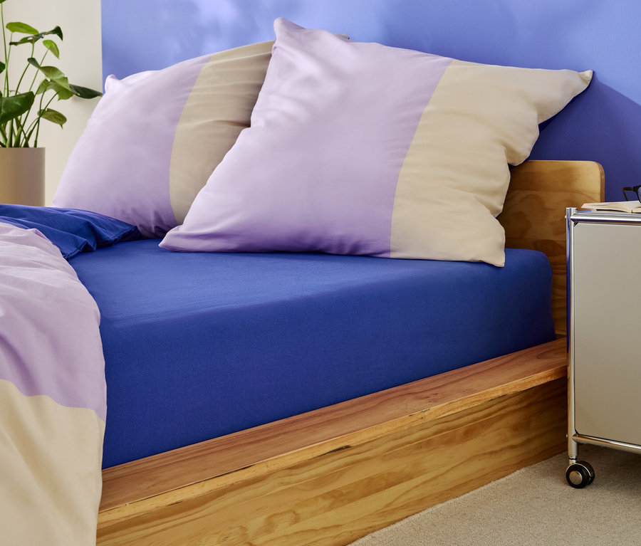 Bett mit blauem Spannbettlaken und Kissen mit lila und beige Streifen.