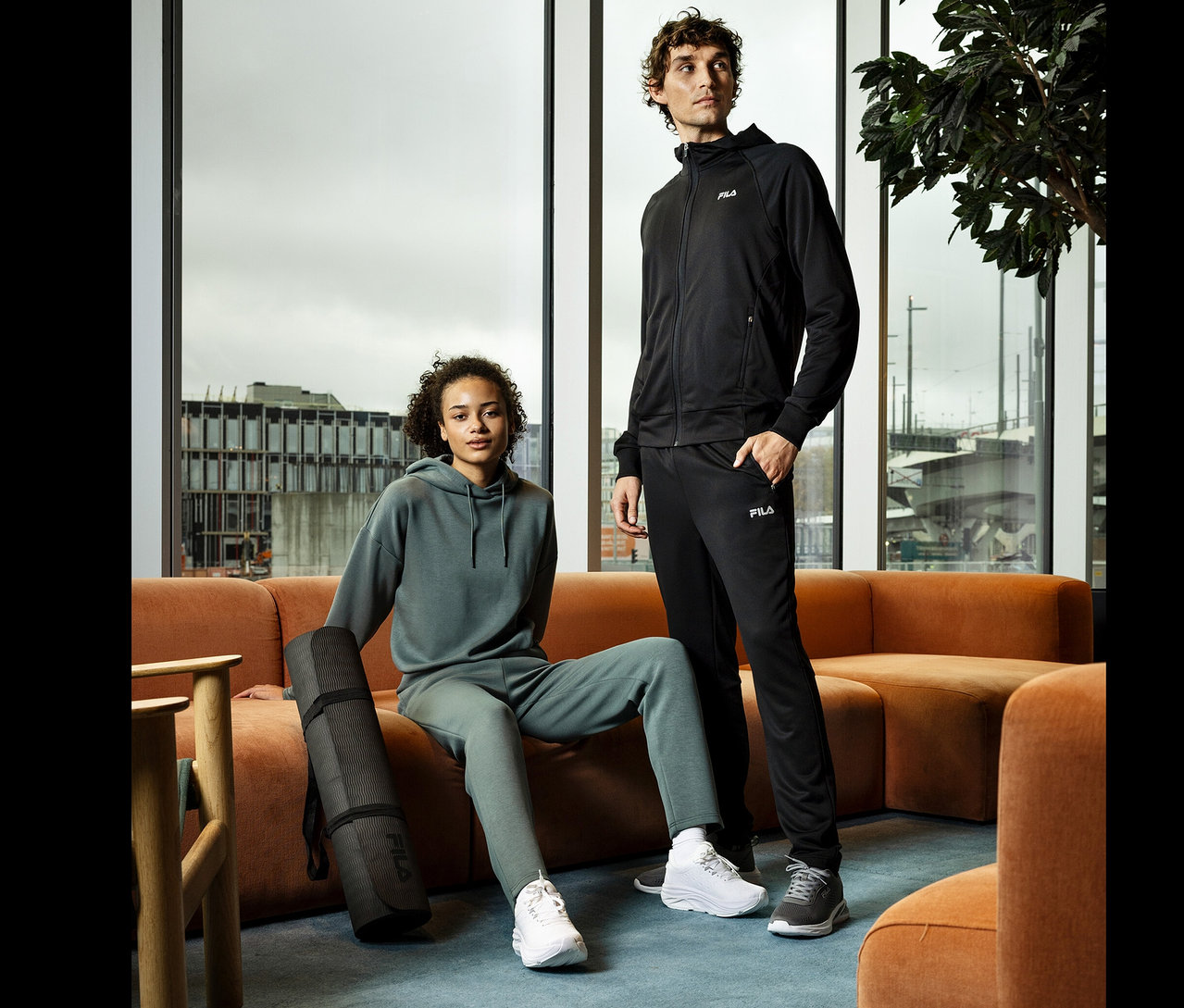 Eine Frau sitzt auf einem orangefarbenen Sofa mit einer schwarzen Yogamatte. Ein Mann steht daneben. Beide tragen Sportbekleidung von Fila.