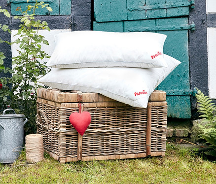 Zwei weiße Paradies 3-Kammer-Daunenkissen »Eimsbüttel« liegen auf einem Weidenkorb mit einem roten Herzanhänger davor.