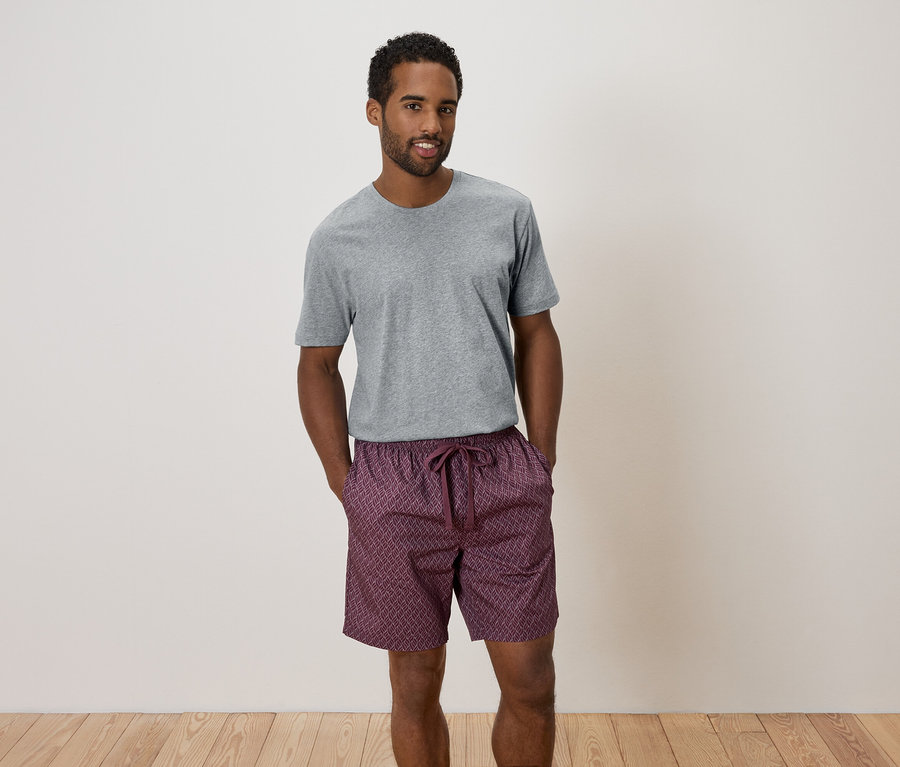 Mann steht mit verschränkten Händen in grauer Hose und gemusterten Shorts da.