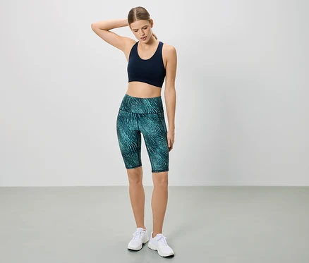 Frau in Sportbekleidung posiert vor einem weißen Hintergrund. Sie trägt einen dunkelblauen Sport-BH und kurze, gemusterte Leggings.