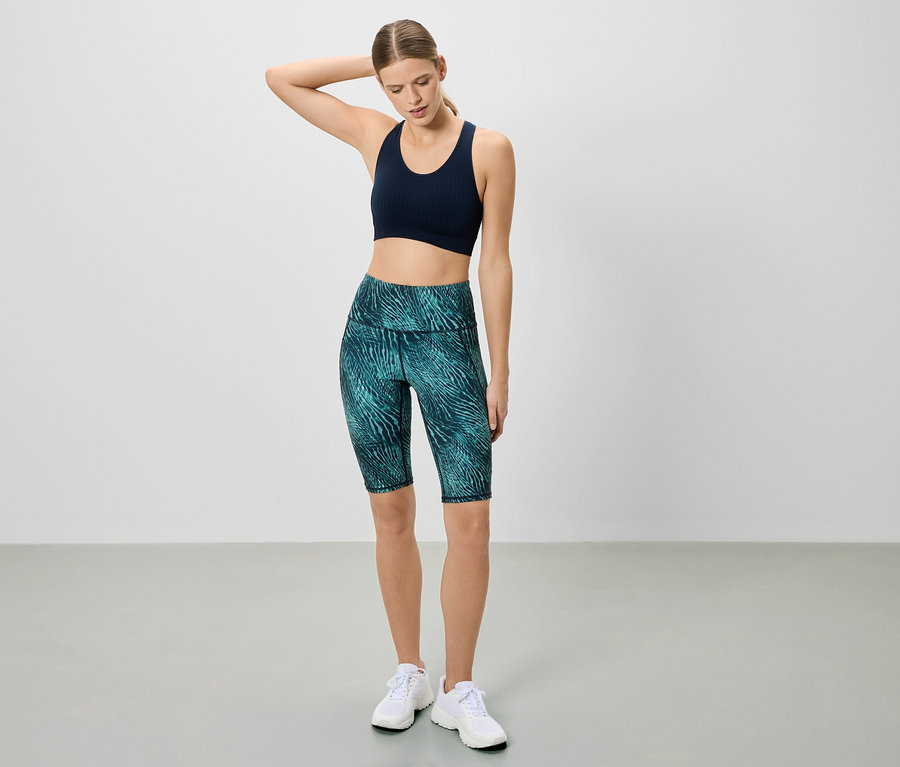 Frau in Sportbekleidung posiert vor einem weißen Hintergrund. Sie trägt einen dunkelblauen Sport-BH und kurze, gemusterte Leggings.