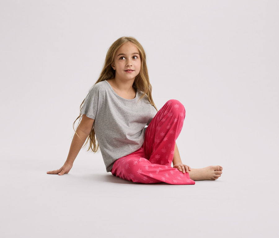Mädchen sitzt auf dem Boden, trägt ein graues T-Shirt und eine pinkfarbene Hose mit Palmenmuster.