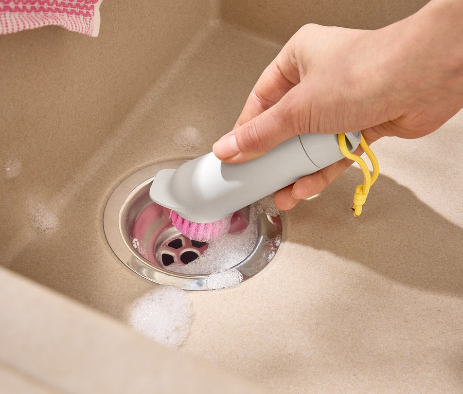 Eine Hand reinigt einen beigen Spülbeckenabfluss mit einer grauen Bürste mit rosa Borsten und gelber Schlaufe.