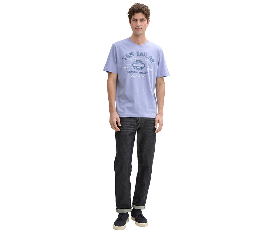 Ein Mann steht in einem hellblauen T-Shirt mit Aufdruck und dunklen Jeans vor einem weißen Hintergrund.