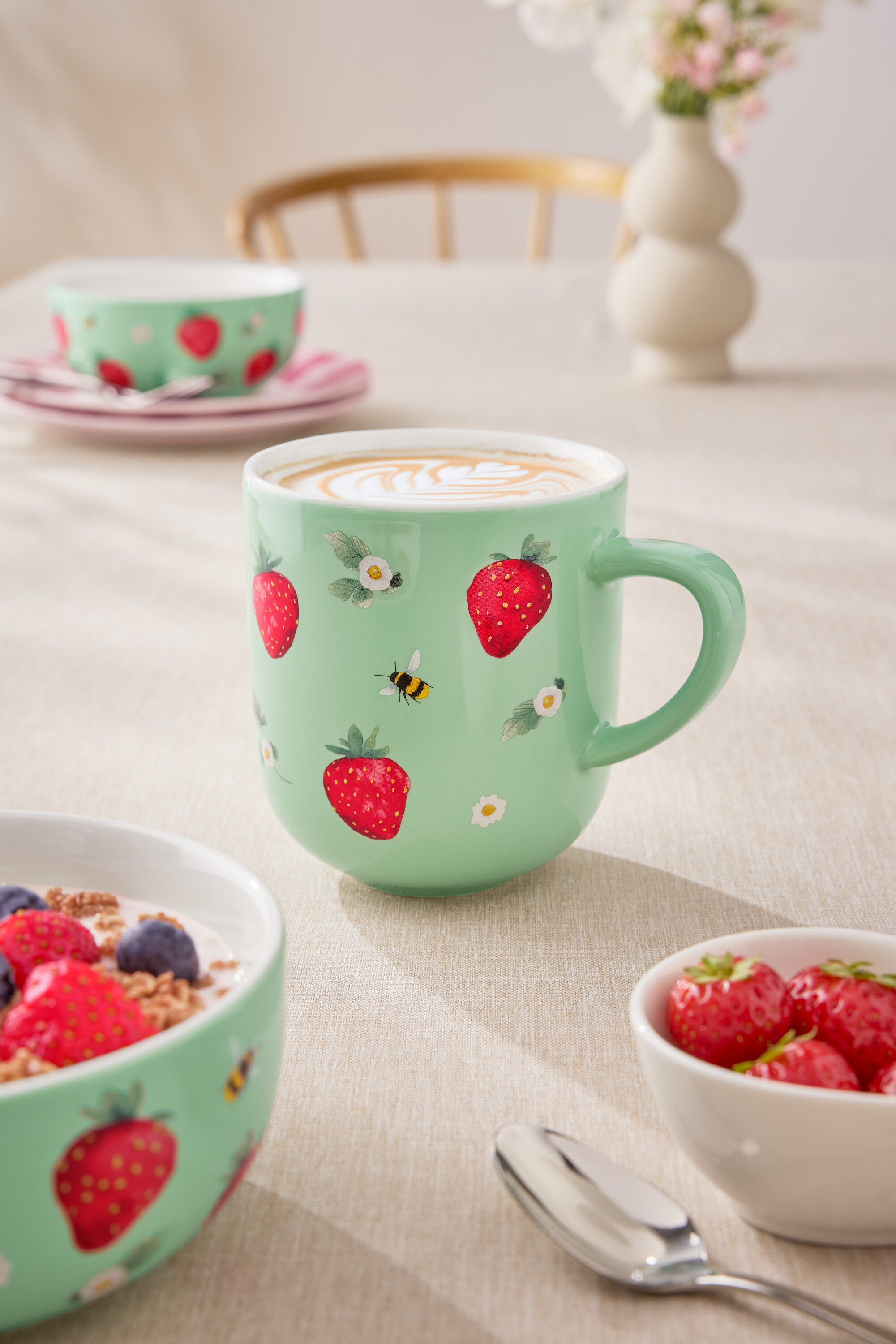 Nahaufnahme eines Tisches mit einer hellgrünen Tasse Kaffee, einer Müslischale mit Joghurt und Beeren sowie einer kleinen Schale mit Erdbeeren. Die Tasse ist mit Erdbeeren und kleinen Blumen verziert.