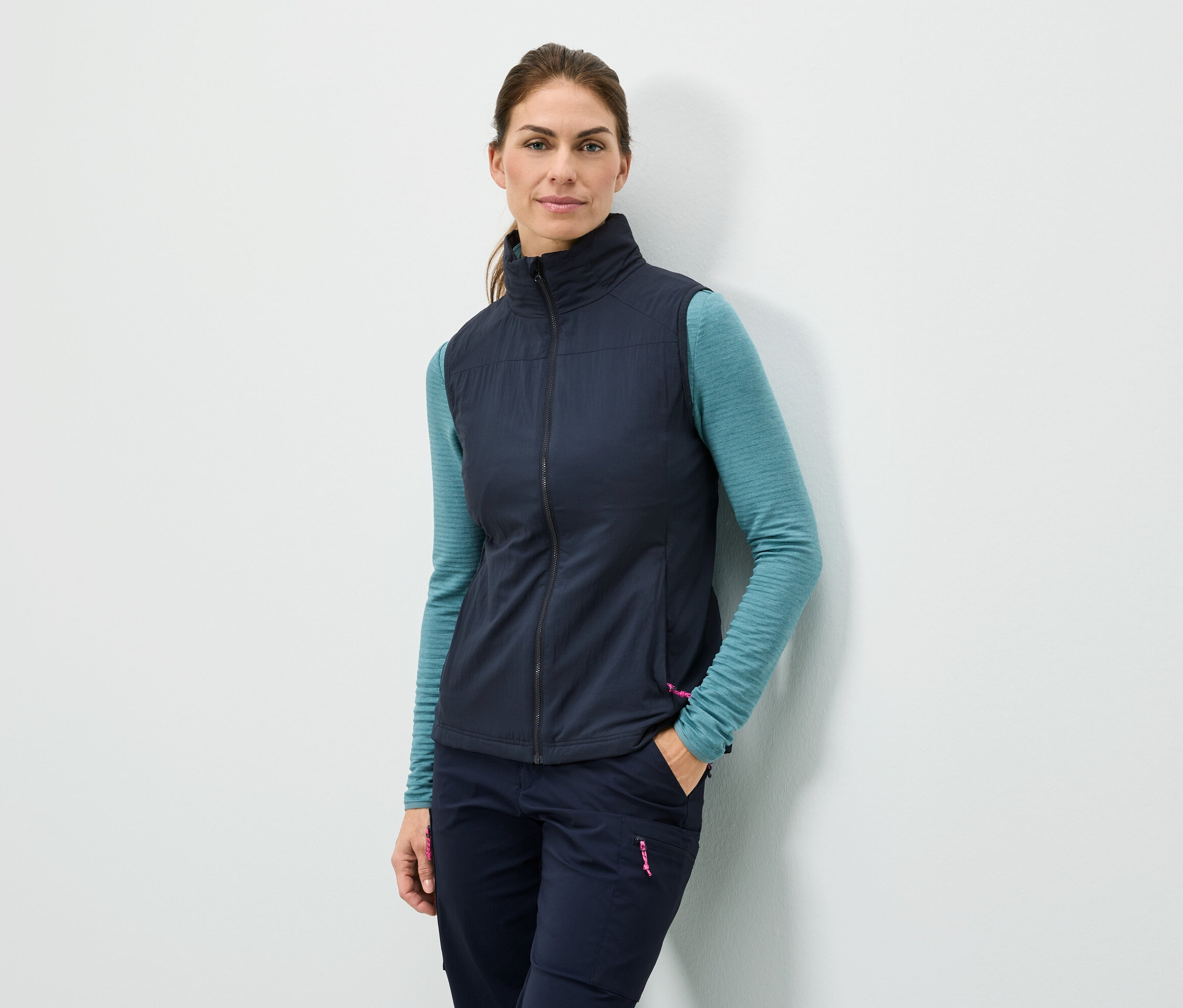 Frau steht mit der Hand in der Tasche an einer Wand, sie trägt eine marineblaue Weste, ein türkisfarbenes Langarmshirt und marineblaue Hosen.