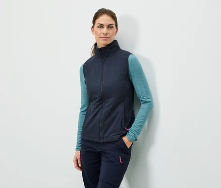 Frau steht mit der Hand in der Tasche an einer Wand, sie trägt eine marineblaue Weste, ein türkisfarbenes Langarmshirt und marineblaue Hosen.
