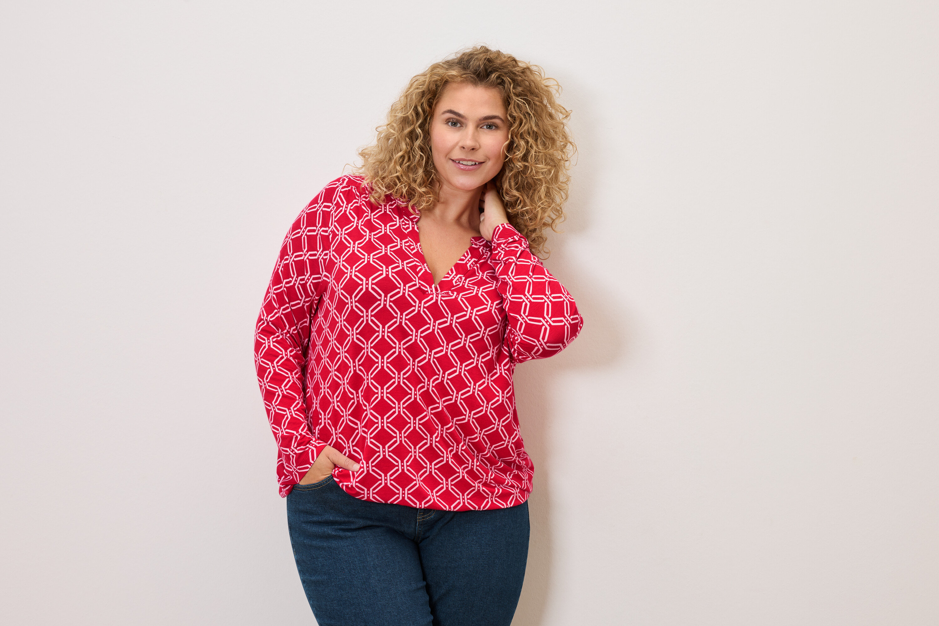 Frau mit lockigen Haaren, die eine rot-weiße Bluse und Jeans trägt und mit einer Hand in der Hosentasche posiert.
