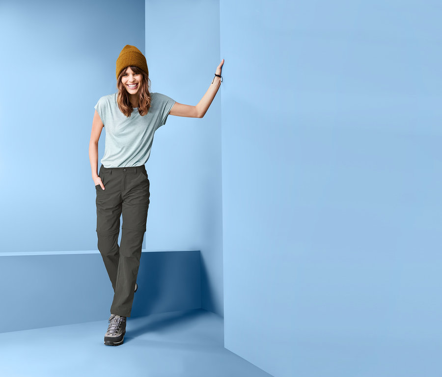 Frau posiert an einer Wand und trägt eine khaki Funktionshose, abzippbar, ein hellblaues T-Shirt und eine braune Mütze.