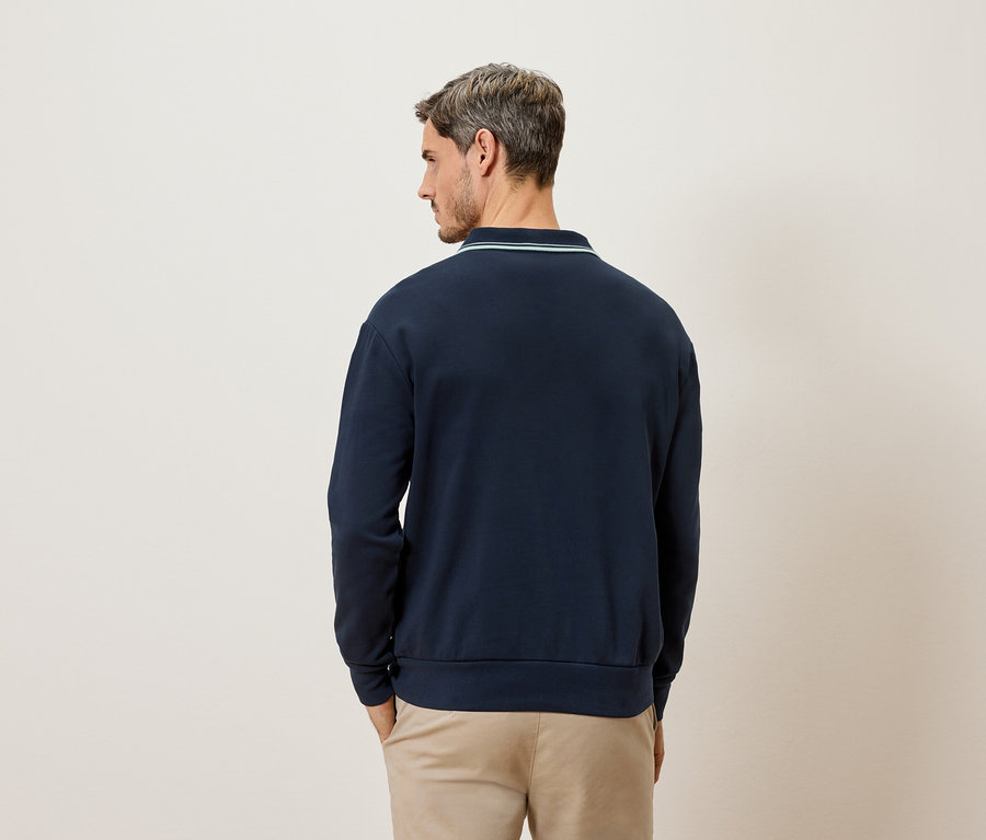 Mann von hinten, trägt ein marineblaues Langarmshirt mit hellgrünem Kragen und beige Hosen.