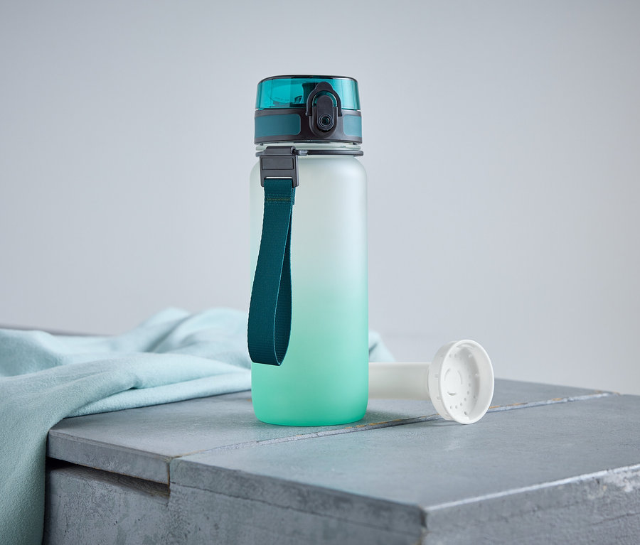 Eine Wasserflasche mit blaugrünem Deckel und Trageband steht auf einer grauen Oberfläche neben einem hellblauen Tuch.