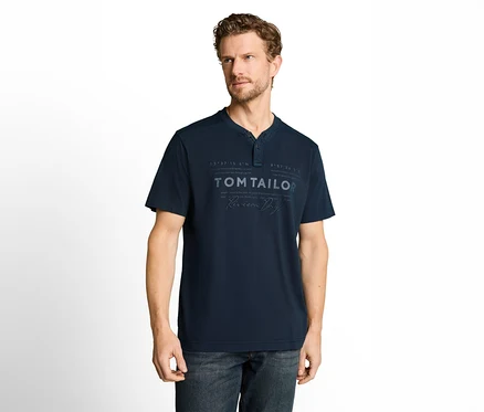 Ein Mann mit lockigem braunem Haar und Bart trägt ein dunkelblaues Kurzarm-T-Shirt mit Henley-Ausschnitt und "TOM TAILOR"-Aufdruck sowie eine dunkle Jeans. Er steht vor weißem Hintergrund und blickt nach rechts.