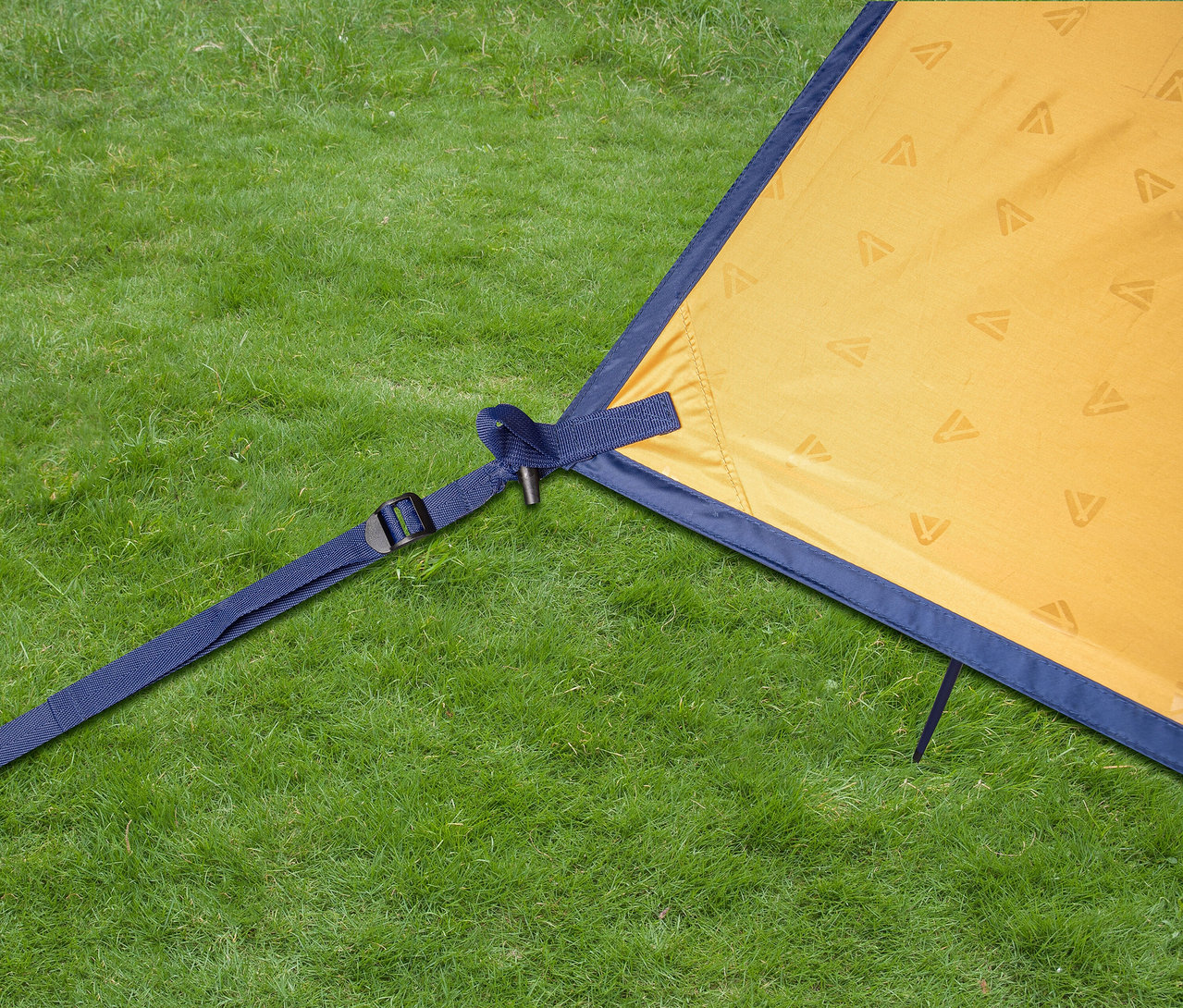 Nahaufnahme eines Tambu »CHADA« Tarps/Sonnensegels auf einer Wiese, befestigt mit einem blauen Band und Hering.