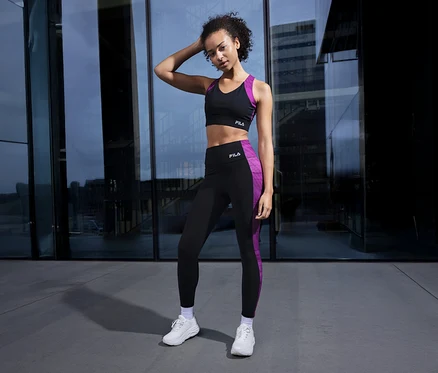 Frau posiert in schwarzem FILA Damen-Sport-BH mit lila Details und passender Leggings vor Glasfassade.