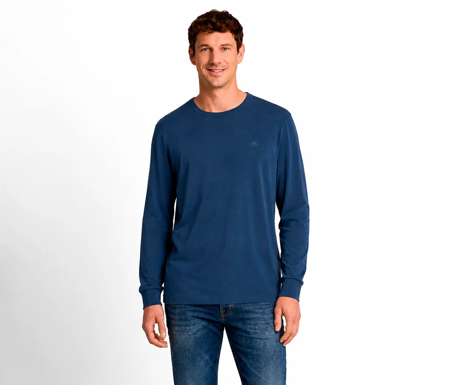 Mann mit kurzen braunen Haaren lächelt, trägt ein blaues Langarmshirt mit kleinem Logo auf der Brust und blaue Jeans, steht vor weißem Hintergrund.