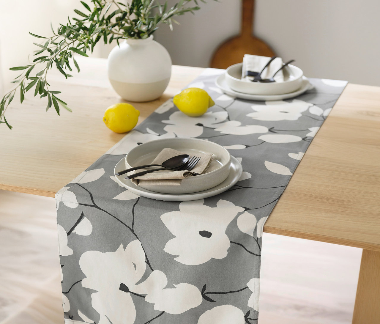 Grauer Tischläufer der SCHÖNER WOHNEN-Kollektion mit weißem Blumenmuster auf einem Holztisch, darauf zwei Gedecke, Zitronen und eine Vase mit Olivenzweigen.