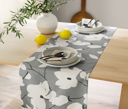 Grauer Tischläufer der SCHÖNER WOHNEN-Kollektion mit weißem Blumenmuster auf einem Holztisch, darauf zwei Gedecke, Zitronen und eine Vase mit Olivenzweigen.