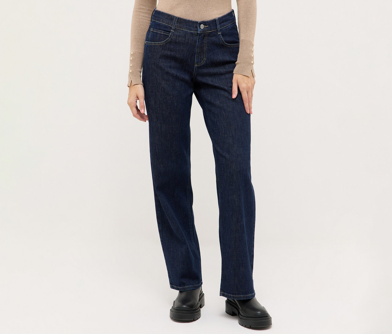 Person trägt dunkelblaue Jeans mit geradem Bein, ein beiges Langarmshirt mit Knopfdetails an den Ärmelbündchen und schwarze klobige Stiefel.