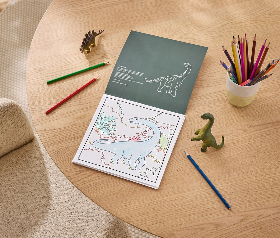 Ein Malbuch liegt auf einem Holztisch und zeigt einen teilweise ausgemalten blauen Dinosaurier. Daneben stehen ein Becher mit Buntstiften, lose rote, grüne und blaue Stifte sowie zwei Dinosaurier-Spielzeuge.