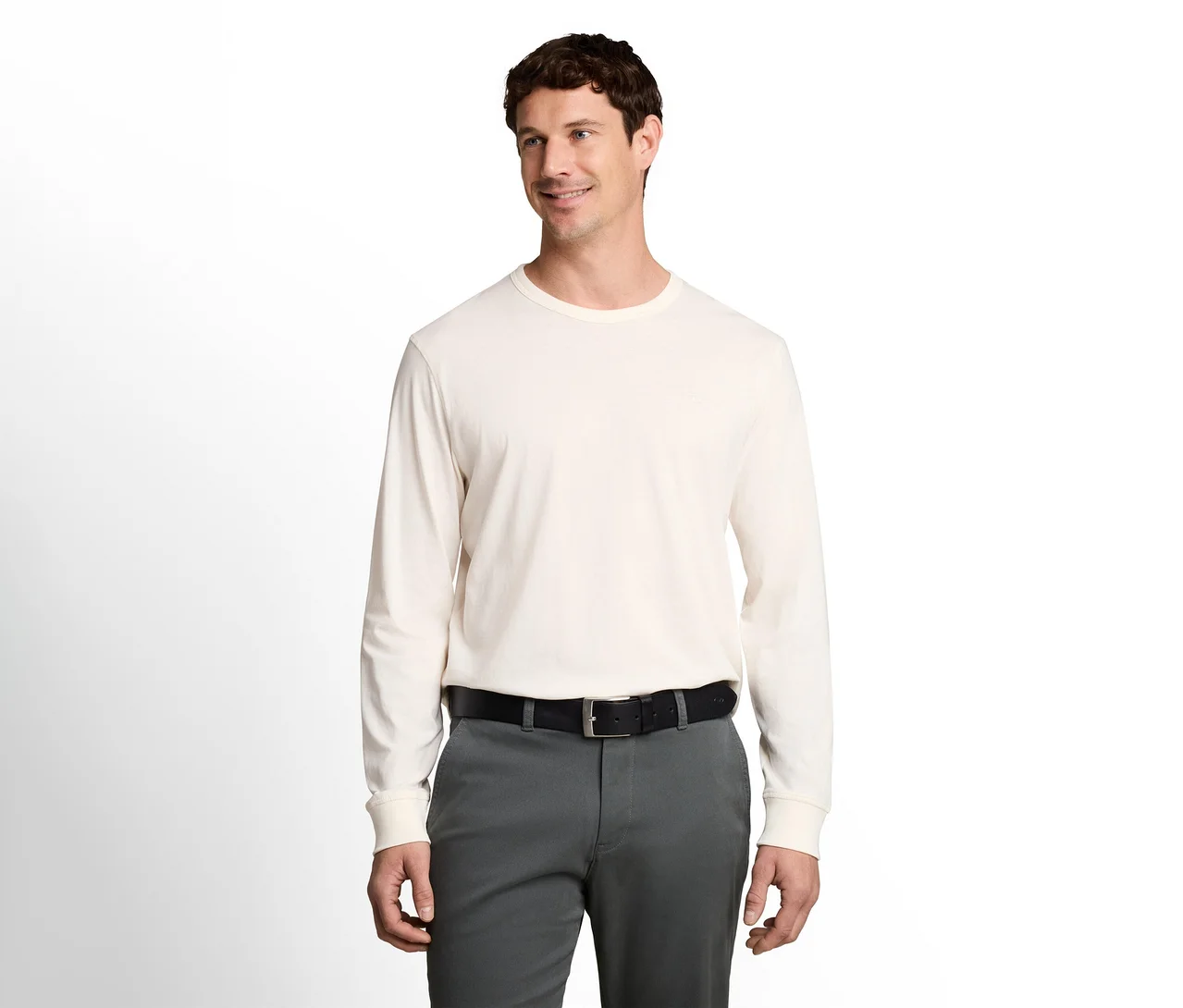 Mann in einem cremefarbenen Langarmshirt, dunkelgrauer Hose und schwarzem Gürtel, lächelt und blickt nach rechts.
