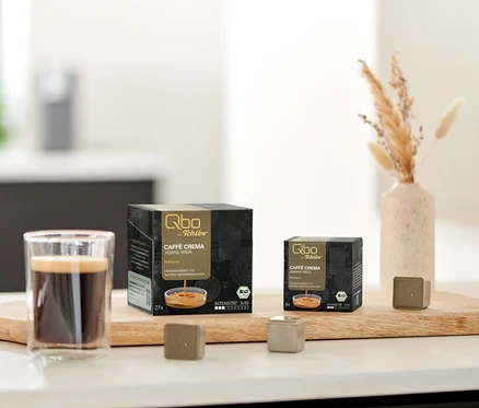Auf einem Brett stehen eine Tasse Kaffee, eine Packung Qbo Caffè JABANA YIRGA - 27 Kapseln und eine Packung Qbo Caffè JABANA YIRGA - 8 Kapseln.