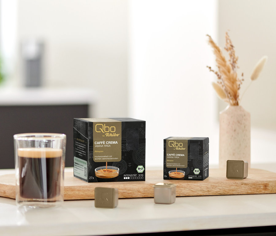 Auf einem Brett stehen eine Tasse Kaffee, eine Packung Qbo Caffè JABANA YIRGA - 27 Kapseln und eine Packung Qbo Caffè JABANA YIRGA - 8 Kapseln.