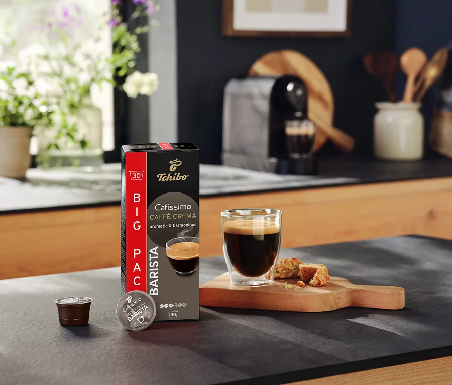 Auf einer Küchentheke stehen eine Packung Tchibo Cafissimo Caffè Crema, eine Kapsel, eine Tasse Kaffee und Gebäck.