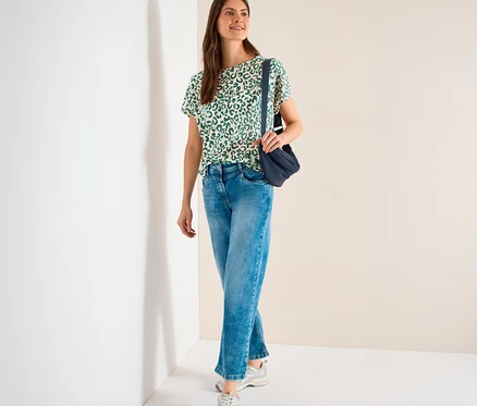 Eine Frau steht lächelnd und trägt ein beiges CECIL Kurzarmshirt mit grünem Leopardenmuster, blaue Jeans, silberne Sneaker und eine dunkelblaue Umhängetasche.