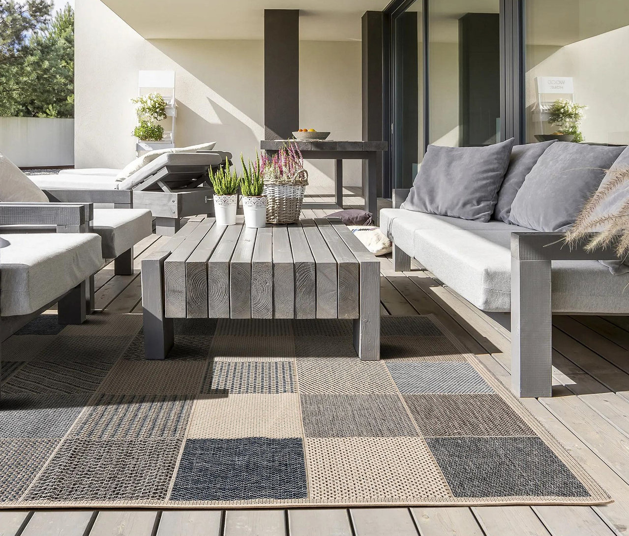 Eine Terrasse mit grauen Holzmöbeln, bestehend aus einem Sofa, einem Sessel und einer Liege mit hellgrauen Polstern, die um einen passenden Couchtisch angeordnet sind. Ein großer rechteckiger Outdoor-Teppich mit Patchwork-Muster in Grau-, Beige- und Blautönen bedeckt den Holzboden. Auf dem Couchtisch stehen Topfpflanzen und ein Blumenkorb.