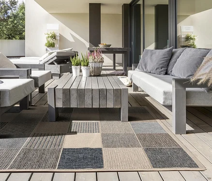 Eine Terrasse mit grauen Holzmöbeln, bestehend aus einem Sofa, einem Sessel und einer Liege mit hellgrauen Polstern, die um einen passenden Couchtisch angeordnet sind. Ein großer rechteckiger Outdoor-Teppich mit Patchwork-Muster in Grau-, Beige- und Blautönen bedeckt den Holzboden. Auf dem Couchtisch stehen Topfpflanzen und ein Blumenkorb.