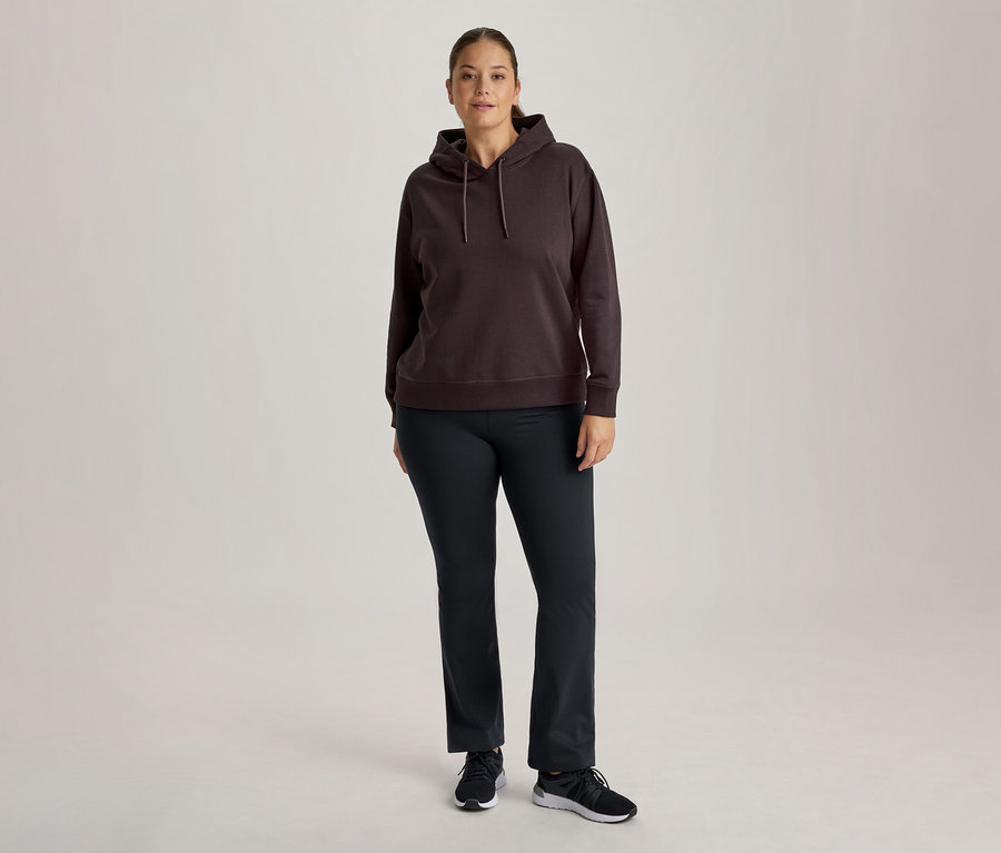Eine Frau steht frontal und trägt einen dunkelbraunen Kapuzenpullover, eine dunkle Hose und schwarze Turnschuhe.