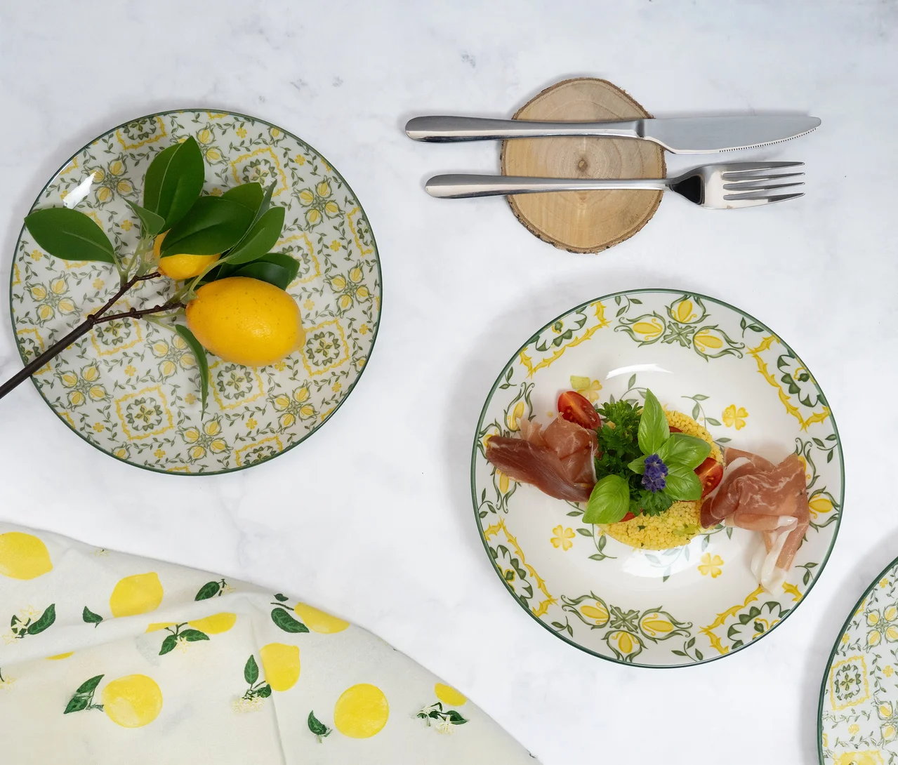Zwei runde Teller mit gelb-grünem Zitronenmuster auf weißem Marmor. Auf einem Teller liegen Zitronen an einem Zweig, auf dem anderen Couscous mit Prosciutto, Basilikum und Tomaten. Daneben liegen ein Messer und eine Gabel des Mäser Besteck-Sets »Ebina« auf einem Holzuntersetzer. Ein Stoff mit Zitronenmuster ist links unten sichtbar.