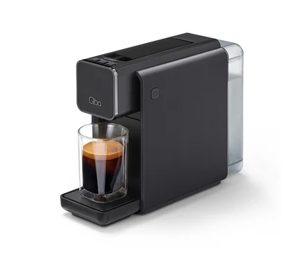 Eine schwarze »Qbo ONE« Kapselmaschine mit einem transparenten Wassertank rechts, die Kaffee in ein kleines Glas auf der Abtropfschale zubereitet.