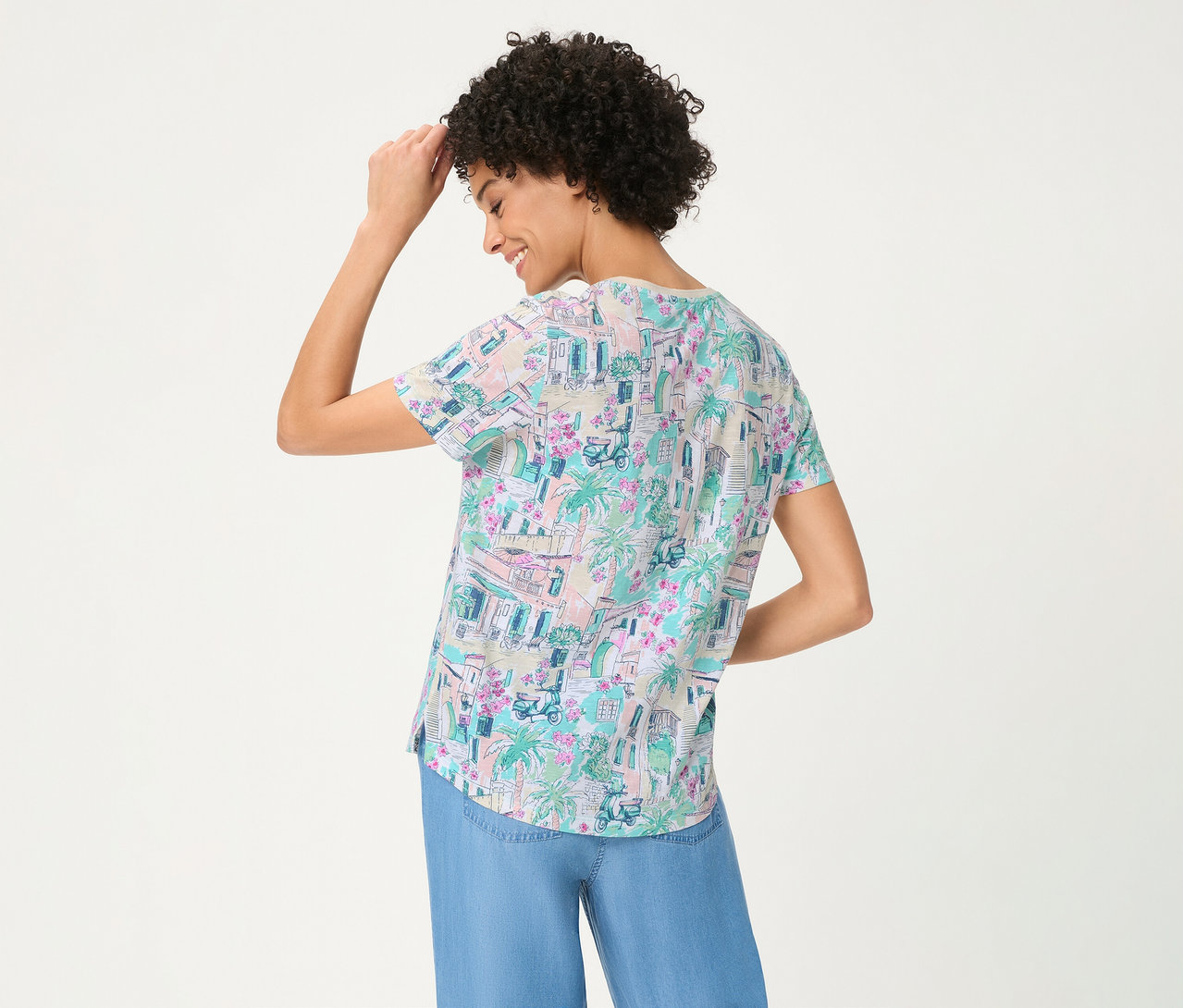 Frau von hinten, lächelt und berührt ihren Kopf, trägt ein Olsen T-Shirt mit buntem Muster aus Gebäuden, Palmen und Rollern, kombiniert mit blauer weiter Hose.