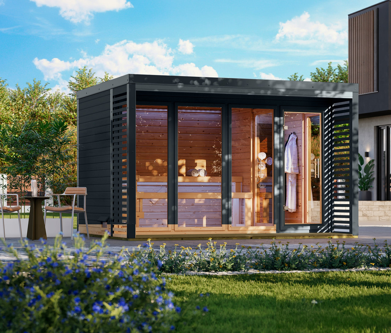 Eine Außensaunakabine mit dunkelgrauer Außenverkleidung und hellem Holzinterieur steht auf einer hellen Terrasse. Die Kabine hat Glastüren, durch die Holzbänke, gerollte Handtücher und ein weißer Bademantel in einem Umkleidebereich sichtbar sind. Links auf der Terrasse stehen ein kleiner dunkler Tisch und zwei Stühle. Im Vordergrund sind grüner Rasen und blaue Blumen zu sehen, im Hintergrund ein modernes Haus unter blauem Himmel.