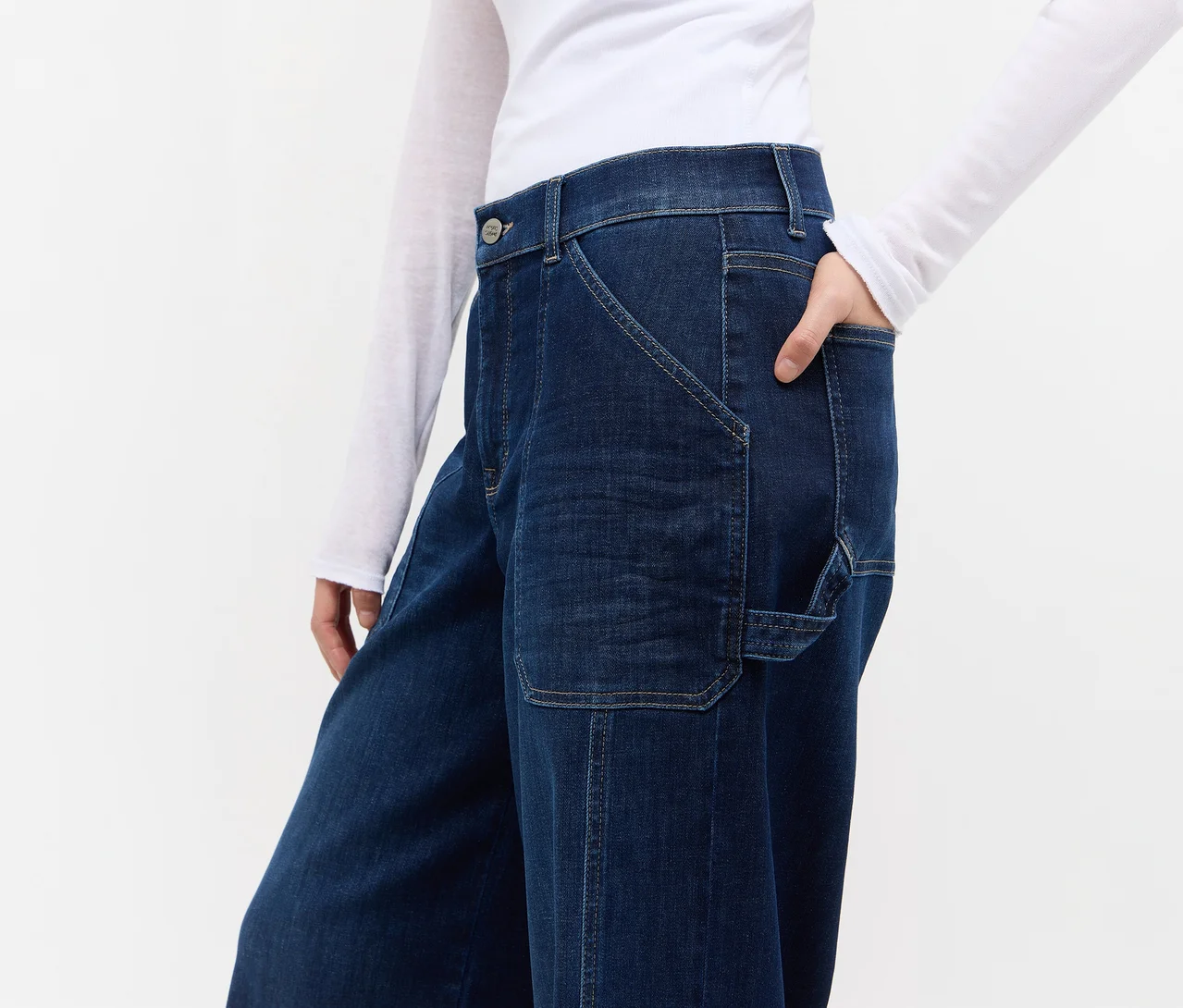 Person in dunkelblauer Jeans mit weitem Bein, aufgesetzten Taschen und sichtbaren Nähten, dazu ein weißes Langarmshirt. Eine Hand steckt in der Tasche.