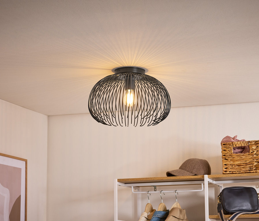 Schwarze Deckenleuchte mit Drahtgeflecht-Schirm, die Licht fächerförmig an die Decke wirft. Darunter ein Regal mit Kleidung, Kappe und Korb.