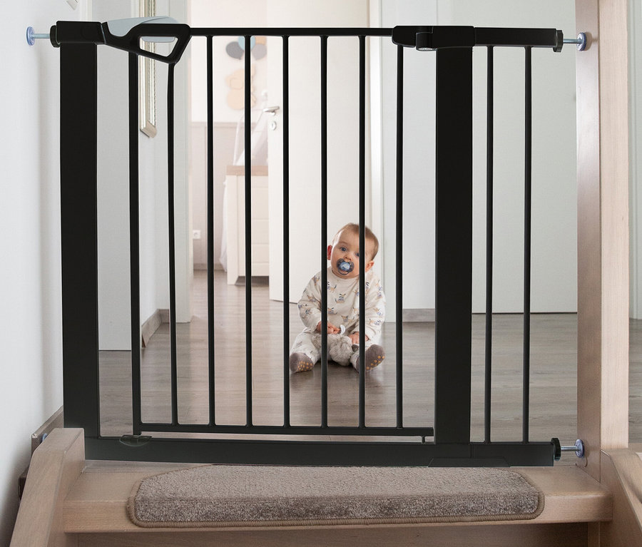 Ein Baby sitzt hinter einem schwarzen roba easySafe+ Tür-und-Treppen-Schutzgitter, das am oberen Ende einer Holztreppe installiert ist. Das Baby trägt einen hellen Strampler und hat einen blauen Schnuller im Mund.