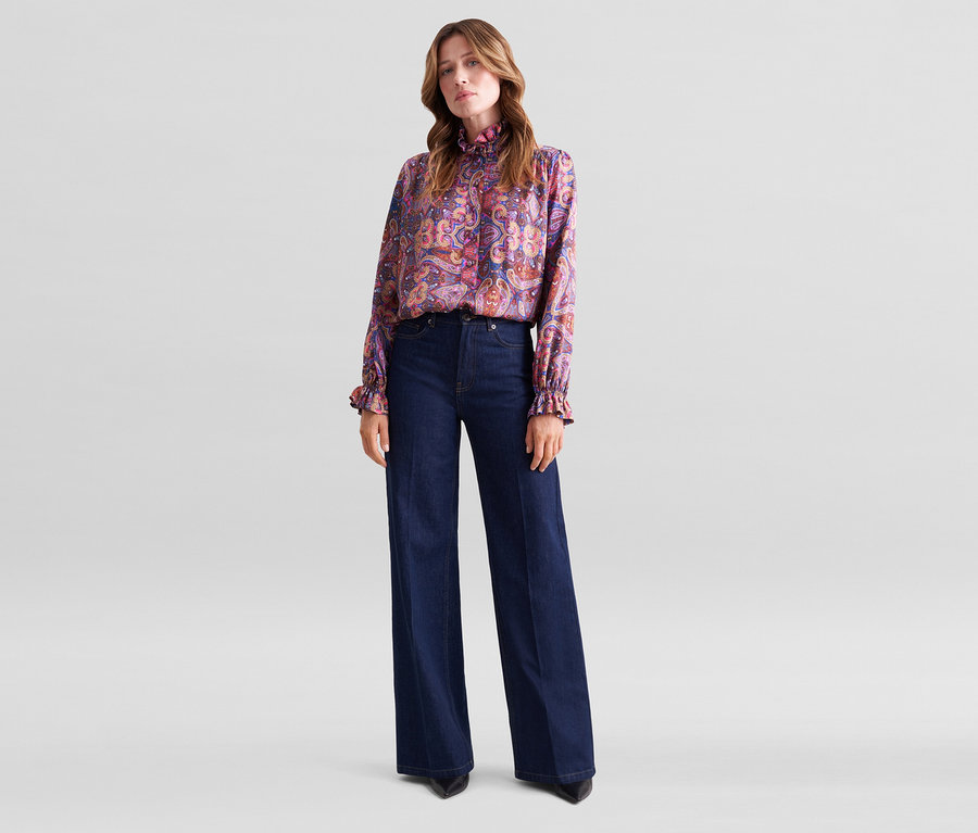 Frau steht in einer bunten Paisley-Bluse mit Rüschenkragen und dunkelblauen weiten Jeans.