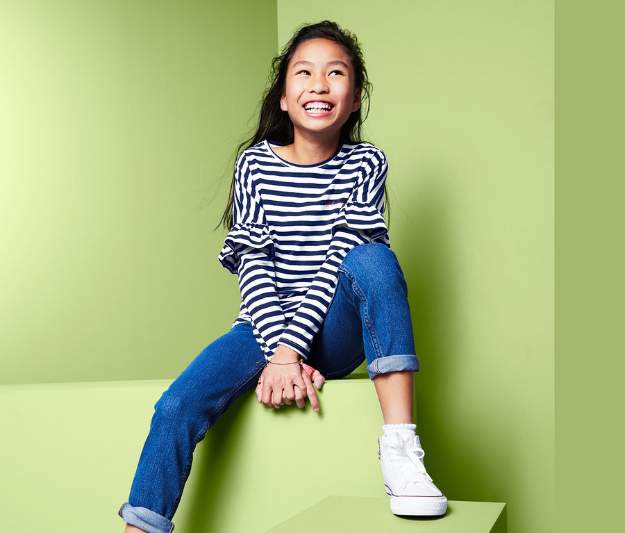 Lächelndes Mädchen sitzt auf grünen Blöcken und trägt ein Langarmshirt mit überschnittenen Ärmeln, Jeans und Turnschuhe.