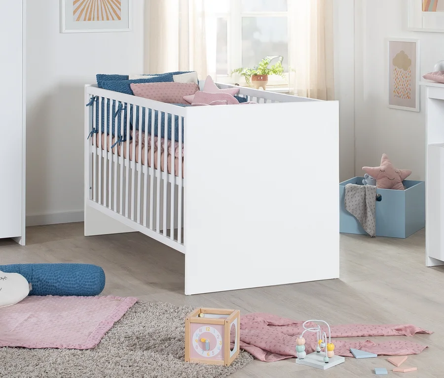 Ein weißes Babybett mit blauen und rosa Kissen sowie rosa Bettwäsche steht auf einem grauen Teppich in einem Kinderzimmer. Daneben befindet sich eine hellblaue Aufbewahrungsbox mit einem rosa Sternenkissen. Auf dem Teppich liegen eine rosa Decke, ein blaues Nackenkissen, ein Aktivitätswürfel, eine Motorikschleife und rosa Babykleidung. Im Hintergrund ist ein Fenster mit einer Pflanze zu sehen.