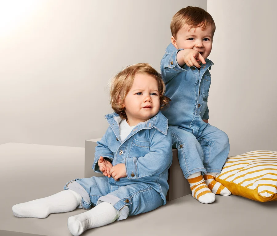 Zwei kleine Kinder posieren vor einem grauen Hintergrund. Beide tragen einen Stretchdenim-Overall.