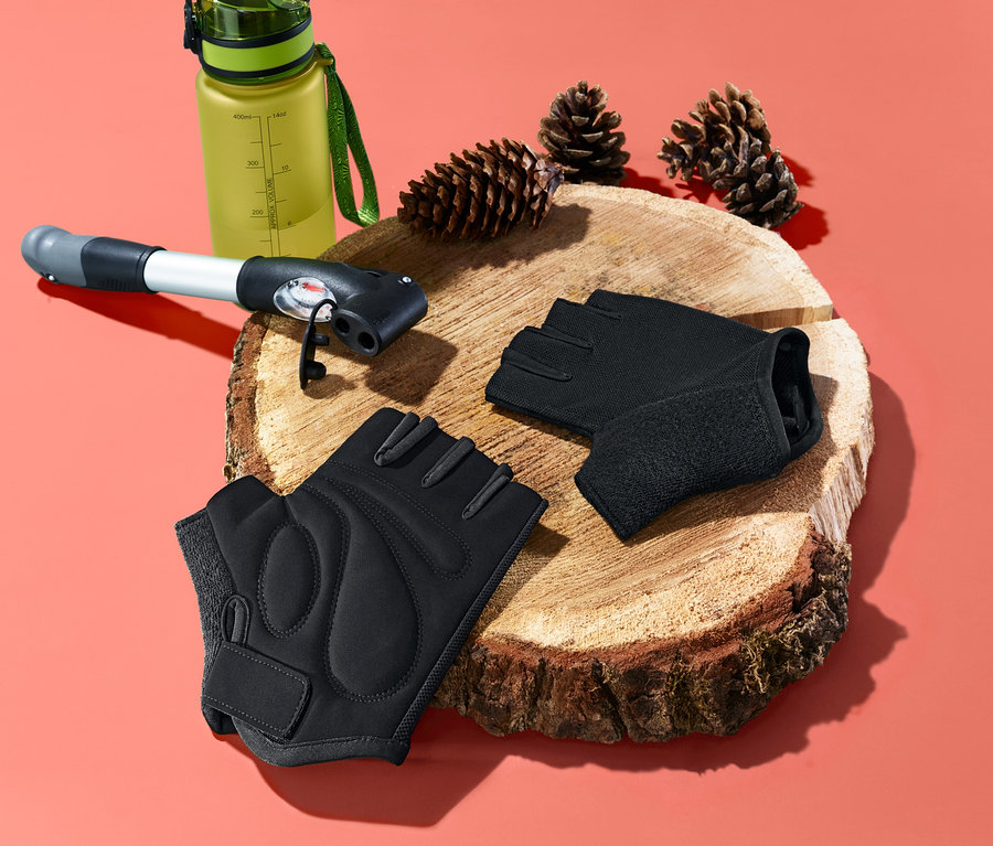 Zwei schwarze Fahrrad-Gel-Handschuhe liegen nebeneinander auf einem Holzbrett. Im Hintergrund befinden sich eine grüne Trinkflasche, eine Pumpe und zwei Tannenzapfen.