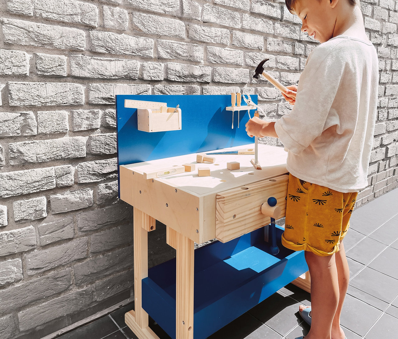 Ein kleiner Junge in hellem Shirt und gelben Shorts spielt mit einer small foot Kinderwerkbank, einen Spielzeughammer über die Holzoberfläche haltend. Die Werkbank ist aus hellem Holz mit blauen Akzenten.