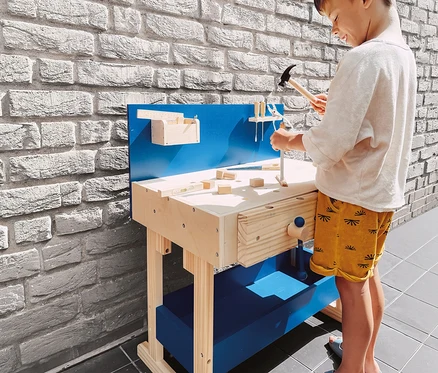 Ein kleiner Junge in hellem Shirt und gelben Shorts spielt mit einer small foot Kinderwerkbank, einen Spielzeughammer über die Holzoberfläche haltend. Die Werkbank ist aus hellem Holz mit blauen Akzenten.