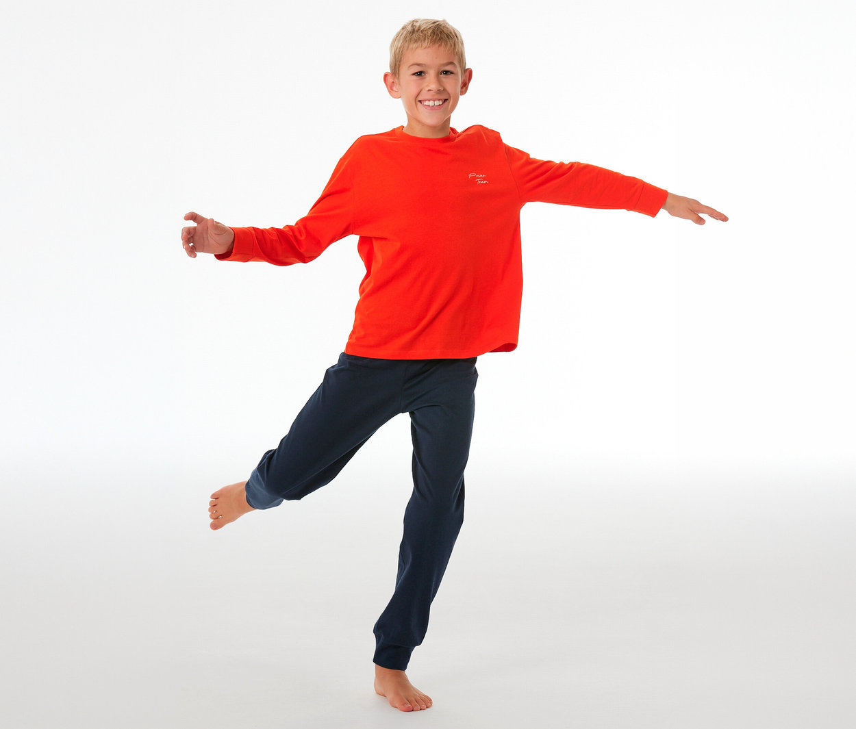 Lächelnder Junge balanciert auf einem Bein, trägt einen SCHIESSER Kinder-Pyjama mit Bündchen: orangefarbenes Langarmshirt und dunkelblaue Hose.